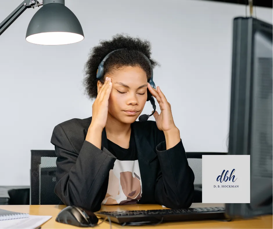 Women at desk holding head with eyes closed.