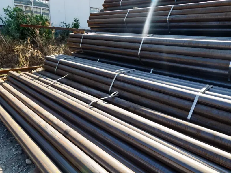 Stacked bundles of dark steel pipes bound with metal straps, stored outdoors near dry brush and a warehouse building.