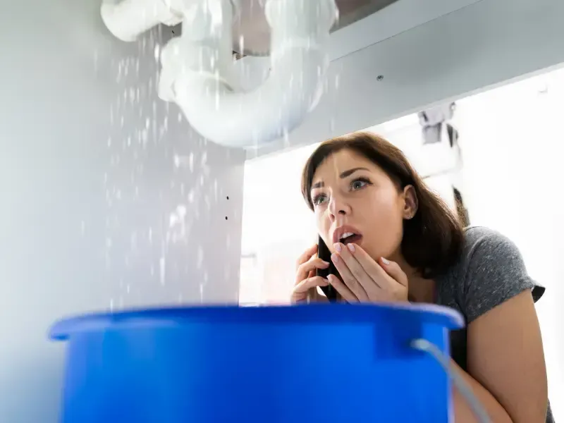 Woman calling a 24/7 emergency plumber while catching a burst pipe leak in a blue bucket.