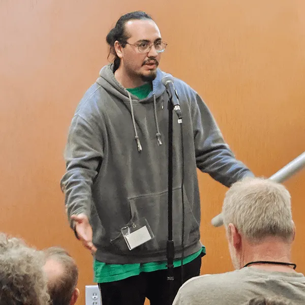 A person in a gray hoodie speaks into a microphone, gesturing to an audience in front of a plain brown wall.