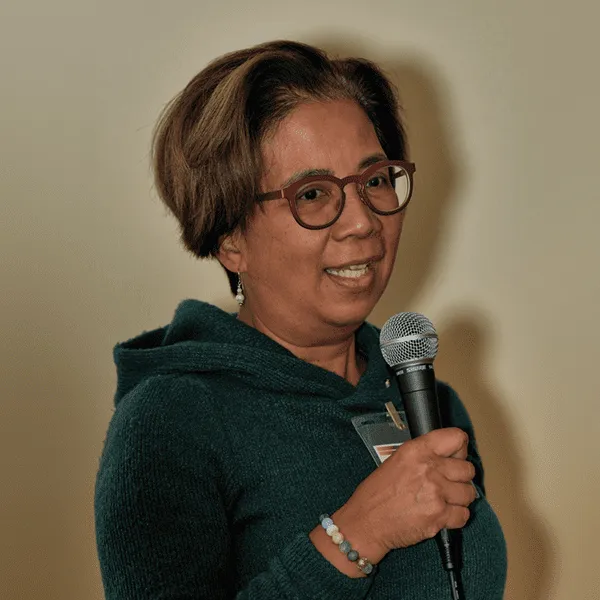 A woman wearing glasses stands confidently, holding a microphone, ready to speak or perform in front of an audience.