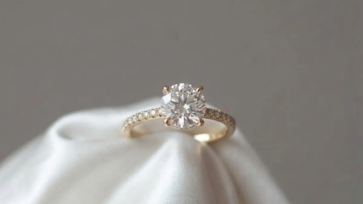 A round brilliant-cut diamond solitaire engagement ring in yellow gold, featuring a delicate pavé band. The ring is elegantly displayed on soft white fabric, highlighting its timeless design, fine craftsmanship, and sparkling center diamond.