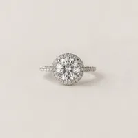 A round brilliant-cut diamond engagement ring featuring a sparkling center stone surrounded by a delicate halo of pavé diamonds. The slim white gold band is adorned with additional pavé-set diamonds, creating an elegant and timeless design. The ring is photographed against a soft neutral background, highlighting its brilliance and refined craftsmanship.