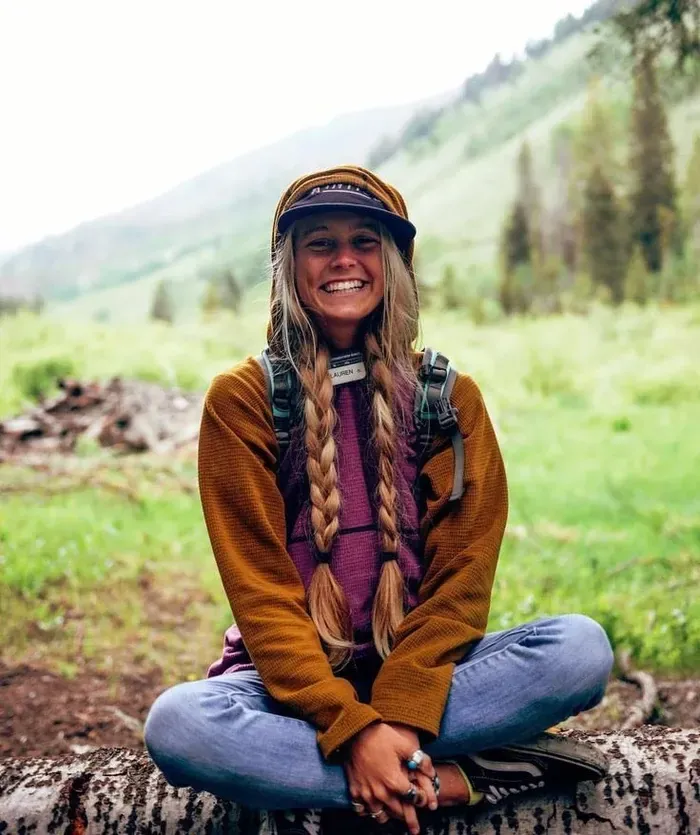 Young woman pictured in nature, sitting cross-legged; beaming smile, blonde hair in messy braids and wearing outdoors clothes appropriate for the nature setting