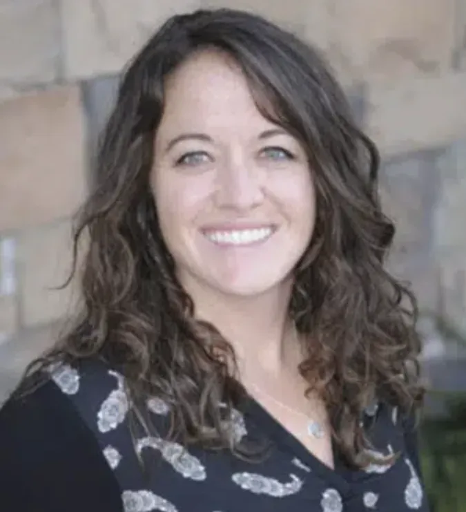 Headshot of Jessica Jo Stenquist, mental health therapist; middle-aged woman with brown curly hair, green eyes, warm smile, with a building backdrop