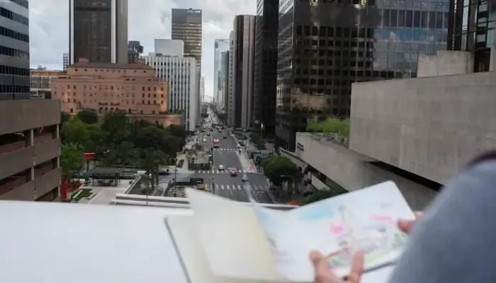 High-angle view looking down a busy city street lined with skyscrapers, with a person in the foreground holding an open sketchbook.