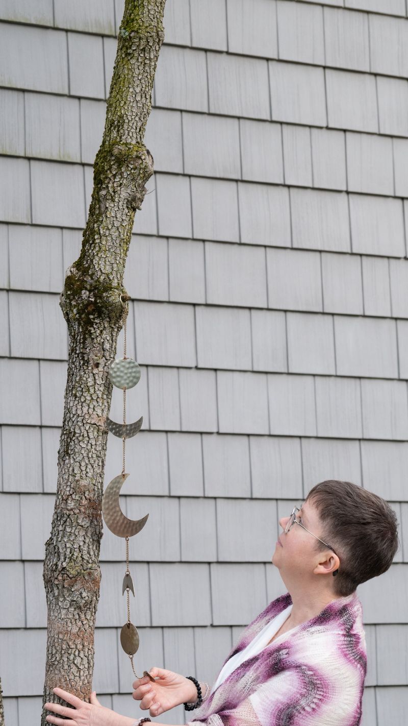 A person with short hair and glasses looks up at a hanging decoration on a tree trunk against a shingled wall, evoking a sense of calm curiosity.
