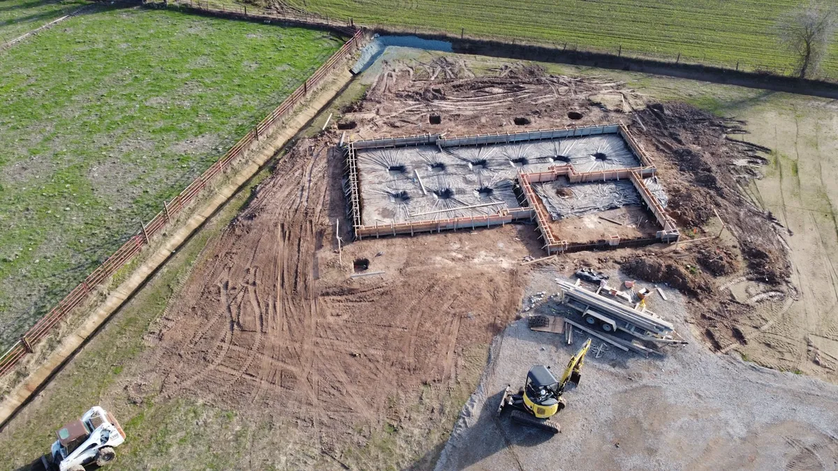 Aerial view of a poured concrete foundation and footings under construction on a graded Central Texas lot, with an excavator and cleared dirt site surrounding the build