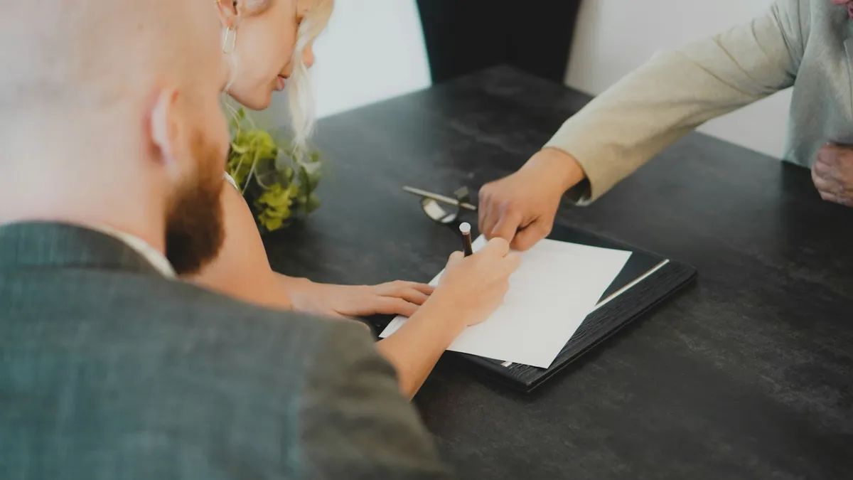 A couple signing a construction contract at a desk, pen in hand, with a project manager pointing to the signature line