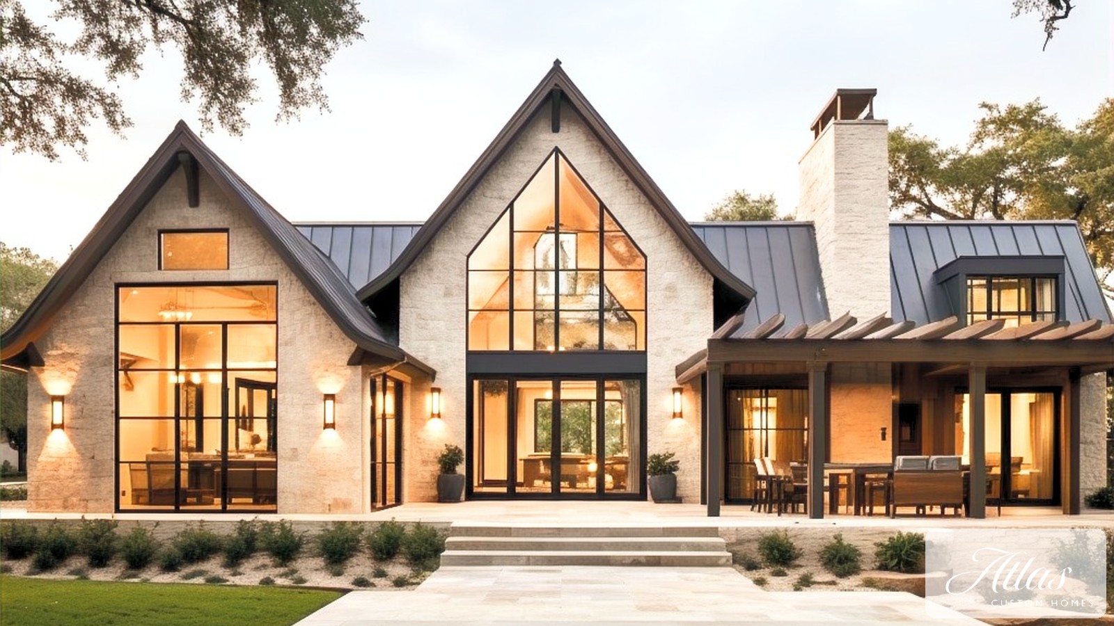 Modern Hill Country custom home with limestone exterior, steep metal roof and glass gable lit at dusk beside a pool, Atlas Custom Homes logo