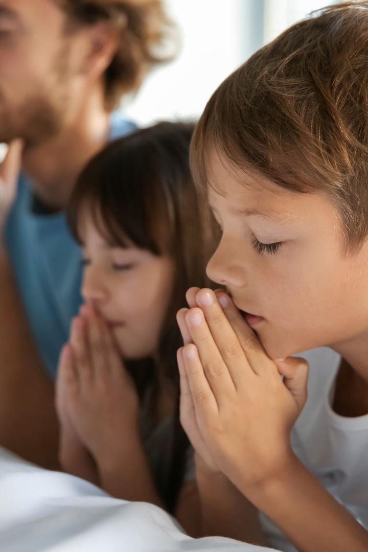 Young children praying with eyes closed and hands clasped alongside a parent, demonstrating family prayer, faith development, and teaching kids the importance of connecting with God.