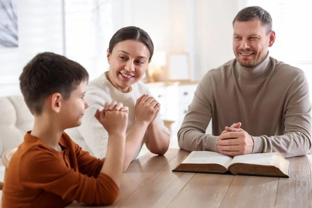 Christian parents and child praying together at the table with an open Bible, demonstrating faith-based parenting, family devotion, and teaching children God’s word at home.