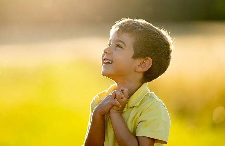 Young boy praying outdoors with hands clasped, smiling with gratitude and faith, representing children learning prayer and trusting God.
