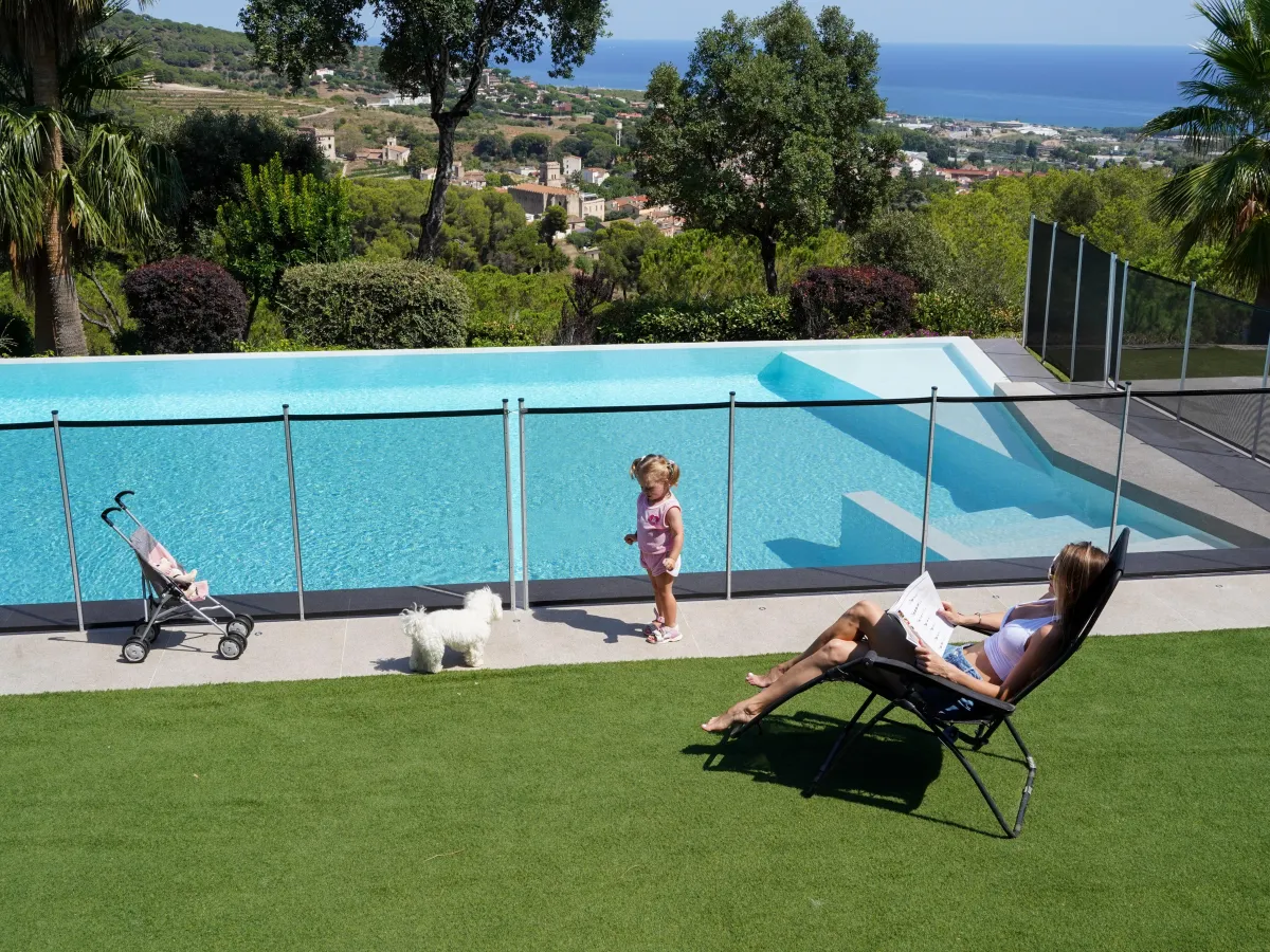 Mujer descansando mientras una niña pequeña y un perro están seguros junto a una piscina protegida con una cerca de seguridad, con vista panorámica al fondo.