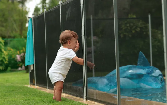 Bebé de pie junto a una cerca de seguridad que protege una piscina, destacando medidas preventivas para evitar accidentes en el hogar.