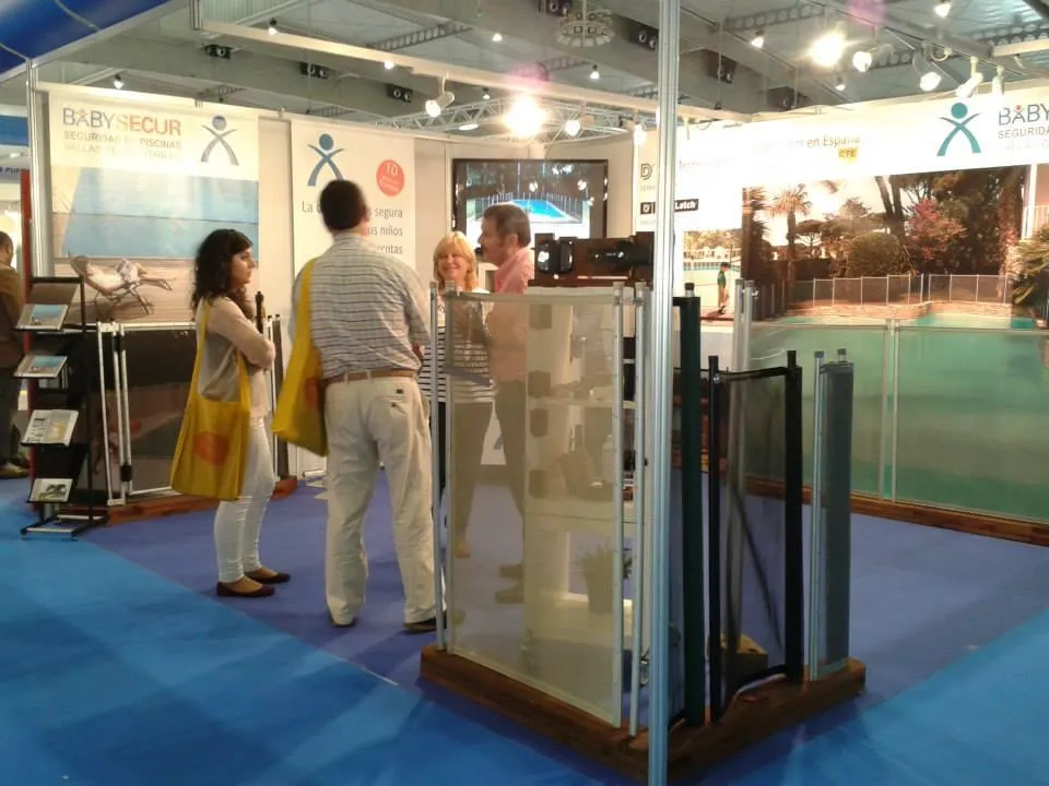  Stand de Babysecur en una feria, mostrando sus vallas de seguridad desmontables y atendiendo a visitantes interesados en soluciones para la protección de piscinas.