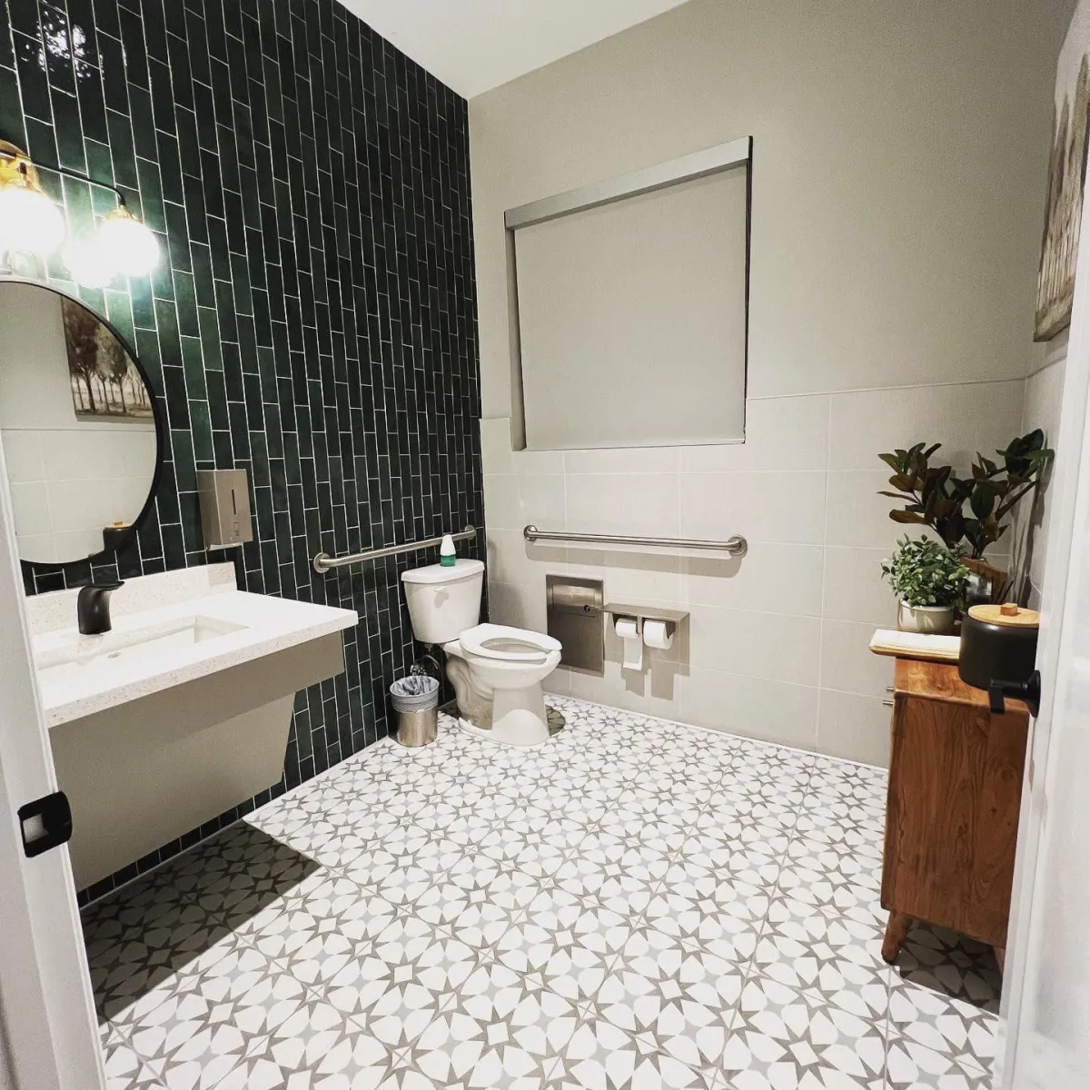 A clean and stylish bathroom featuring a modern design with green subway tiles, patterned flooring, and a sleek white sink. The space includes a well-maintained toilet, accessible grab bars, and a neatly organized wooden cabinet with potted plants, reflecting a fresh and sanitized environment. This showcases **True Cleaning Experts'** attention to detail and commitment to providing spotless, professional cleaning results.