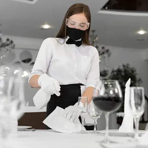 A professional cleaner in a black-and-white uniform with gloves, a face mask, and protective eyewear is sanitizing a formal dining table. The table is elegantly set with wine glasses, napkins, and tableware, highlighting **True Cleaning Experts'** commitment to providing thorough and hygienic cleaning for dining and event spaces.