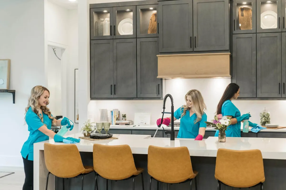 Three professional cleaners from **True Cleaning Experts** are working in a bright, modern kitchen. They are wearing matching blue uniforms and gloves while cleaning various surfaces: one is wiping the countertops, another is cleaning the sink, and the third is sanitizing the stove area. The kitchen features sleek gray cabinets, stylish decor, and a white island with flower arrangements, emphasizing the spotless and organized results delivered by the cleaning team.