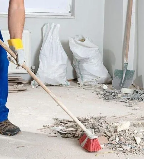 A professional cleaner from **True Cleaning Experts** is sweeping construction debris in a post-renovation setting. The worker, wearing gloves and work attire, uses a sturdy red broom to clean up dust, rubble, and plaster. Large debris bags and tools, like a shovel, are visible in the background, highlighting the team's expertise in **post-construction cleaning services**, ensuring spaces are clean, safe, and move-in ready.