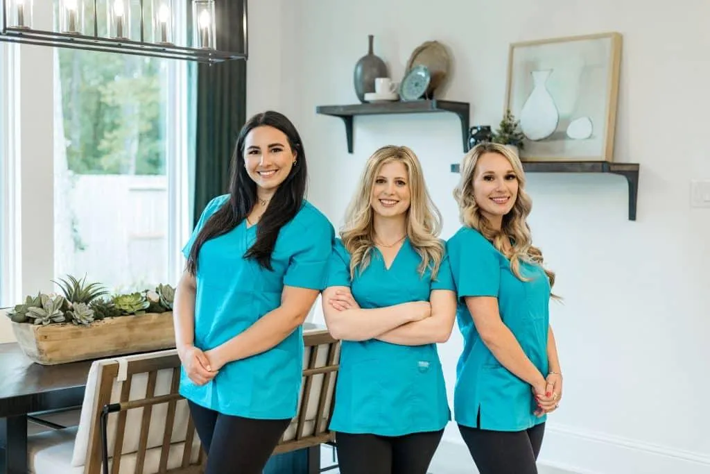 Three professional cleaners from **True Cleaning Experts** are smiling and posing confidently in a modern, clean dining room. They are wearing matching blue uniforms, reflecting their teamwork and professionalism. The background features a bright and organized space with a decorative shelf, framed art, and lush green plants, showcasing a fresh and welcoming environment that highlights their commitment to excellence in cleaning services.