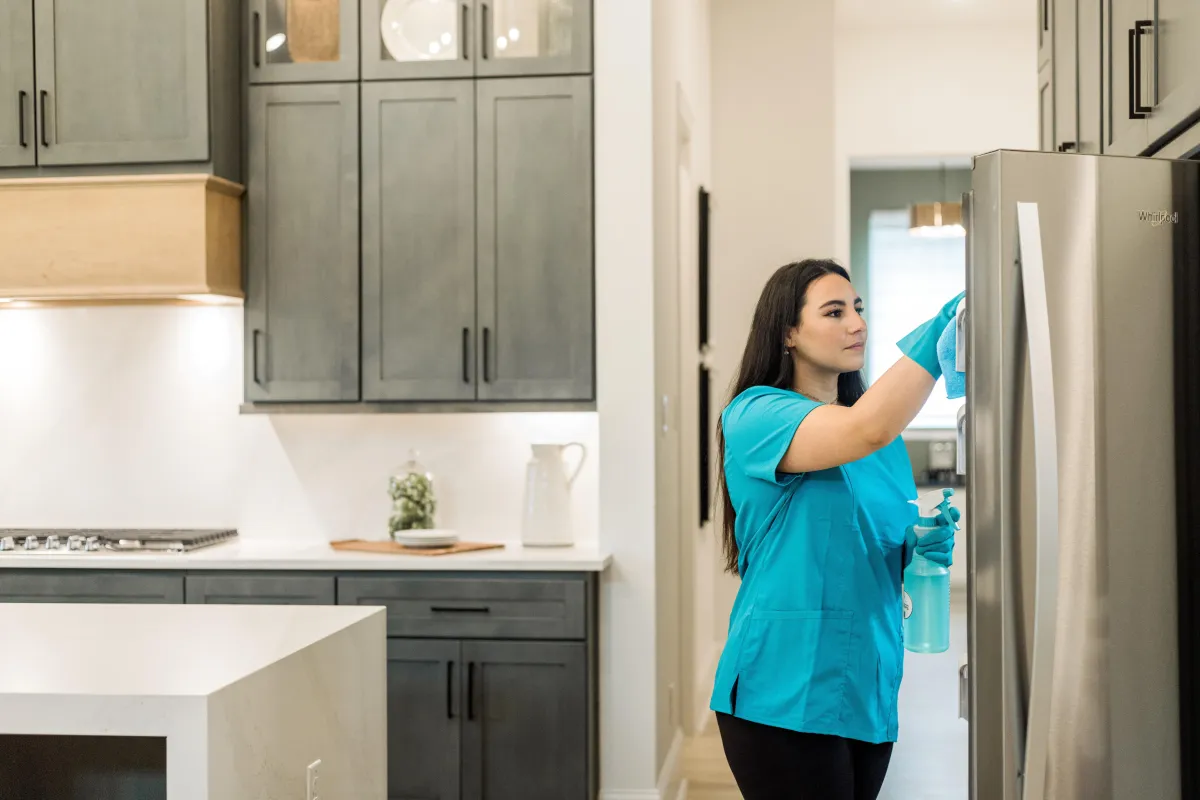 A professional cleaner from **True Cleaning Experts** is wiping down a stainless steel refrigerator in a modern, spotless kitchen. She is wearing a blue uniform and gloves while holding a spray bottle and cleaning cloth. The kitchen features sleek cabinetry, bright lighting, and organized countertops, showcasing the high-quality cleaning standards provided by the team.