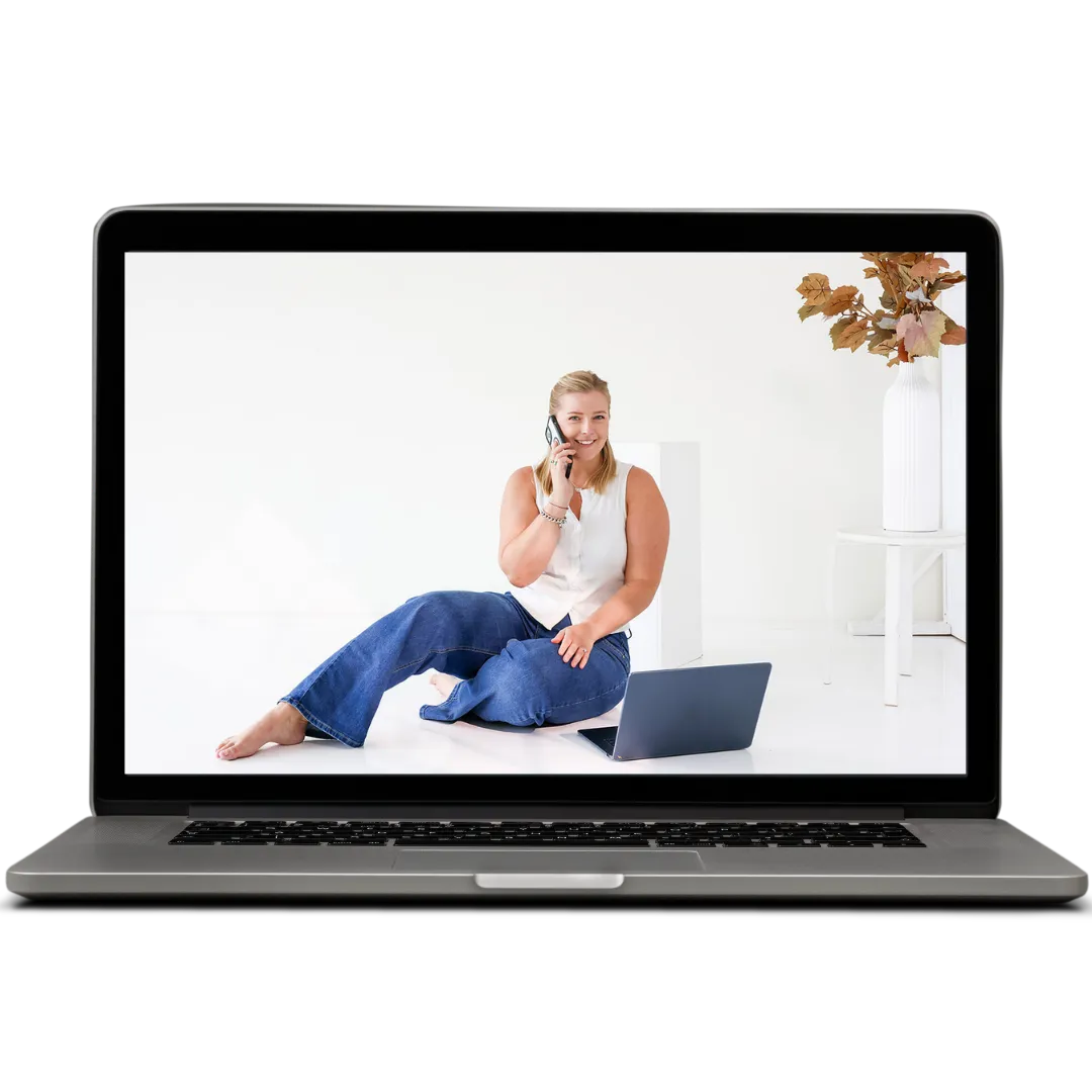 A silver laptop open, displaying a woman sitting on the floor in blue jeans and a white sleeveless top, holding a phone to her ear, smiling. Beside her is another laptop and behind her is a minimalist white setup with a vase containing autumn leaves. The laptop is placed against a stark white background.