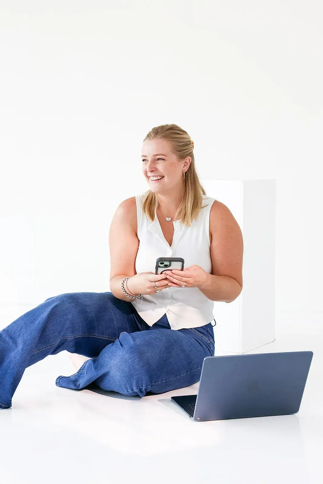 A polaroid-style framed photograph of a woman named Paige sitting casually on a white floor, holding a smartphone, smiling. A laptop is next to her. The photo frame has a small piece of pale yellow tape at the top.