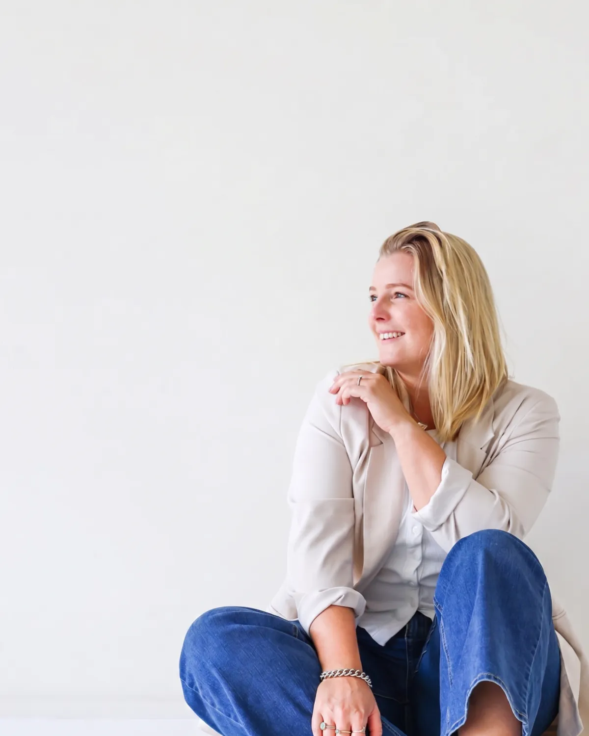 A blonde woman sitting on the floor against a white wall, wearing a beige blazer over a white shirt and blue jeans, looking off to the right side with a slight smile.