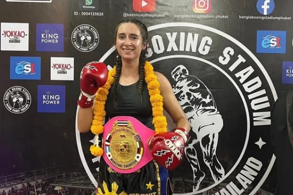Kru Caroline posing with a Muay Thai title belt in Thailand. She had red boxing gloves on and stands before a sponsorship logo board.