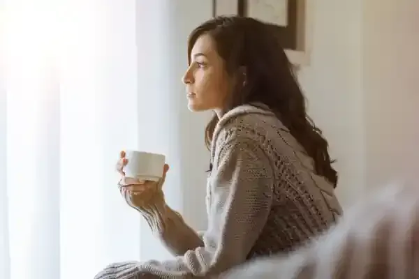 A woman with her hair in a messy bun, wearing a soft beige sweater, sitting by a window holding a white mug