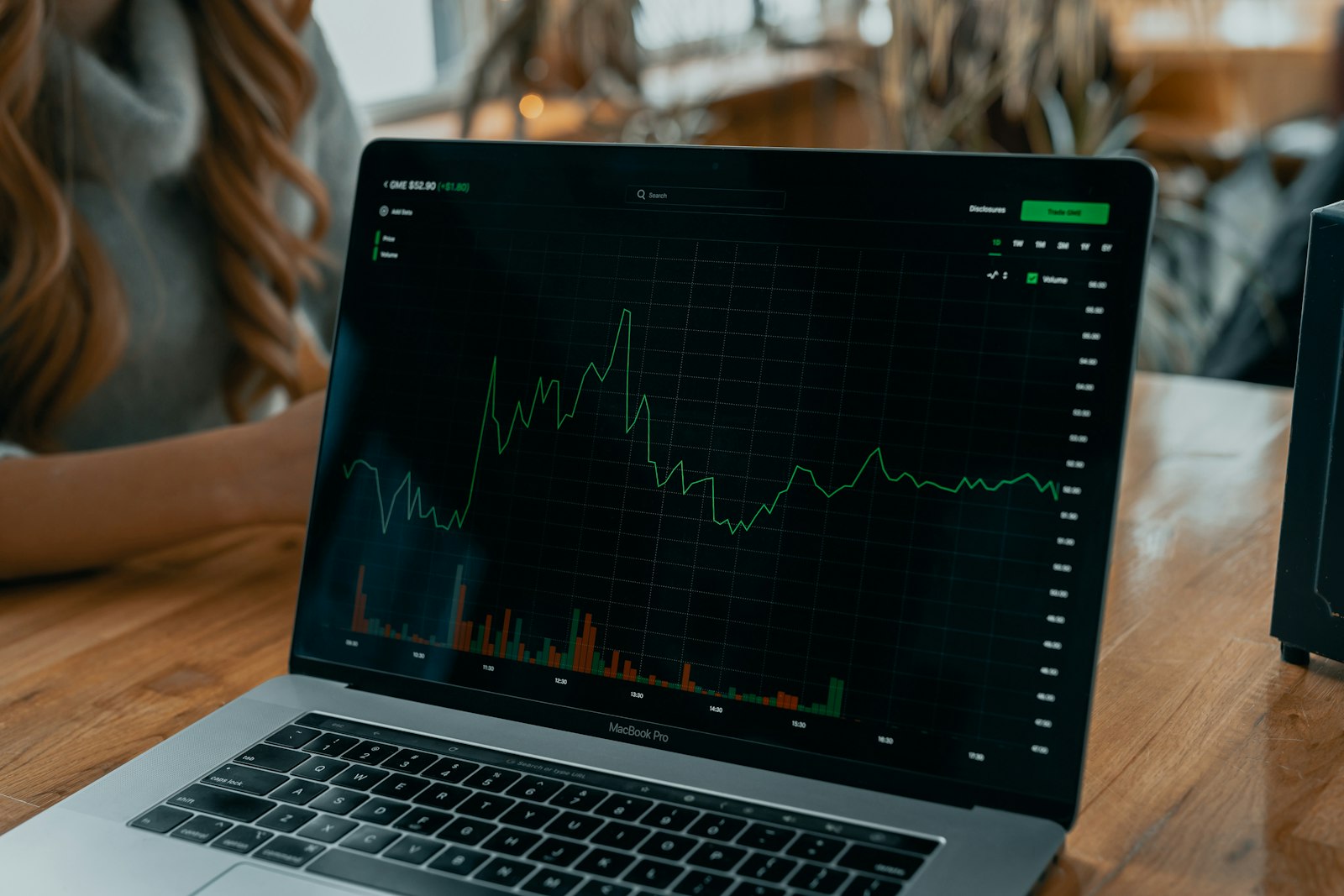 Laptop on a wooden desk displaying a live stock chart in a warm, daylight coffee-shop setting