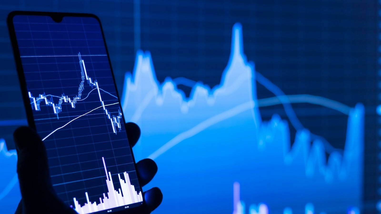 Hand holding a phone showing a stock candlestick chart, with a blurred market index chart glowing in the background