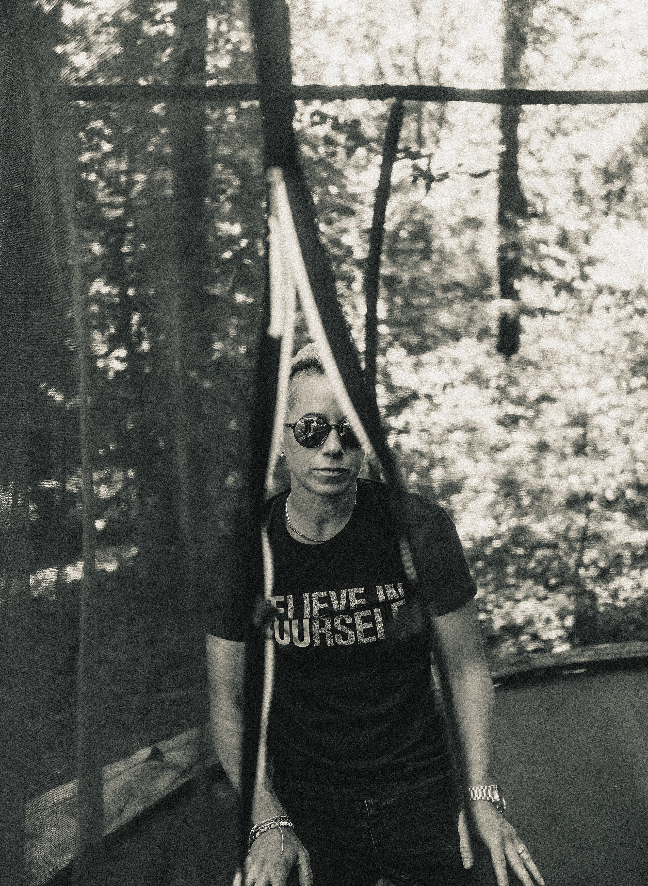 Sepia-toned photo of a woman wearing sunglasses and a t-shirt that says 'BELIEVE YOURSELF', standing in a wooded area.