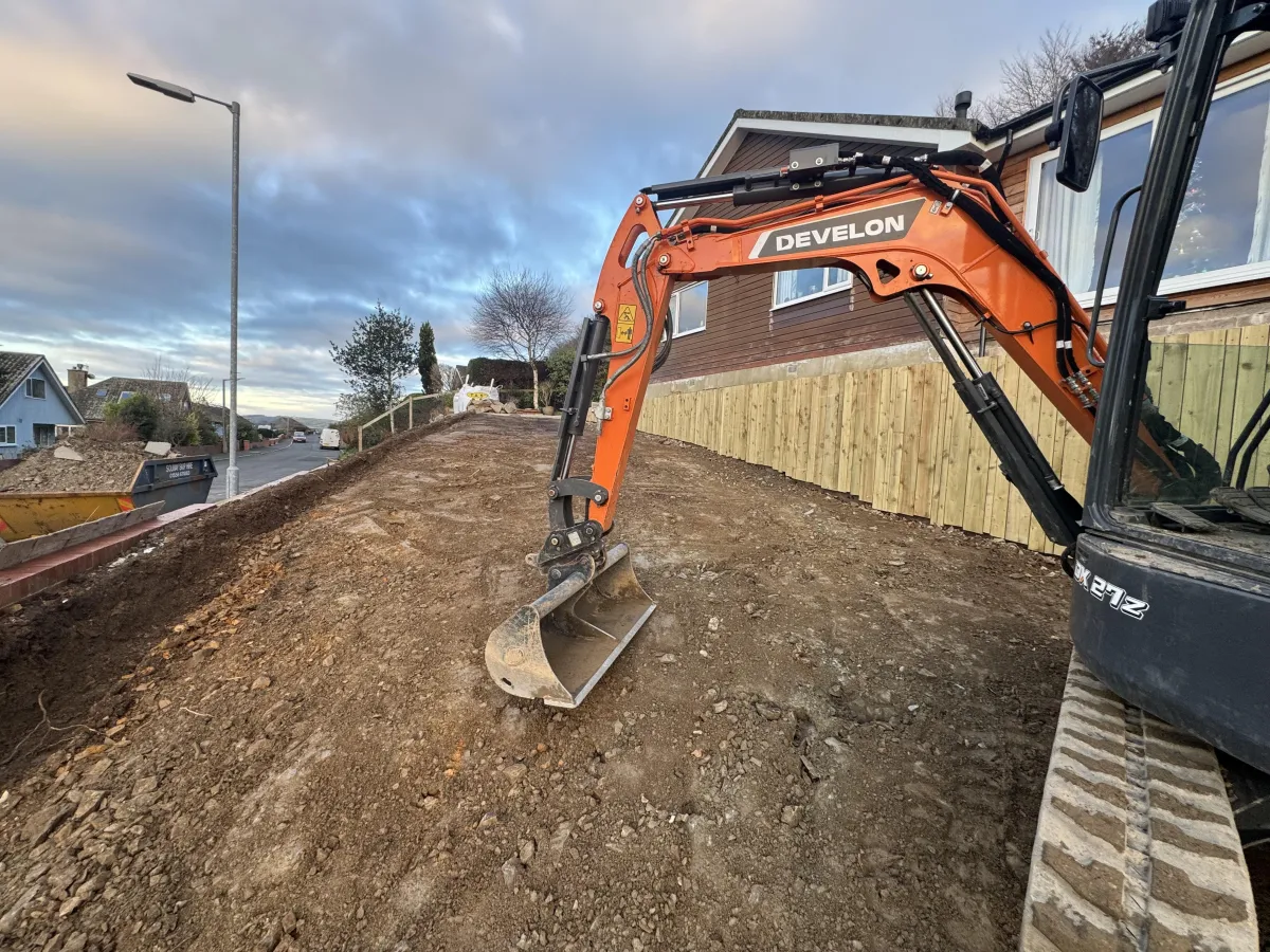 Develon excavator carrying out ground preparation for concrete driveway and foundations beside a residential property