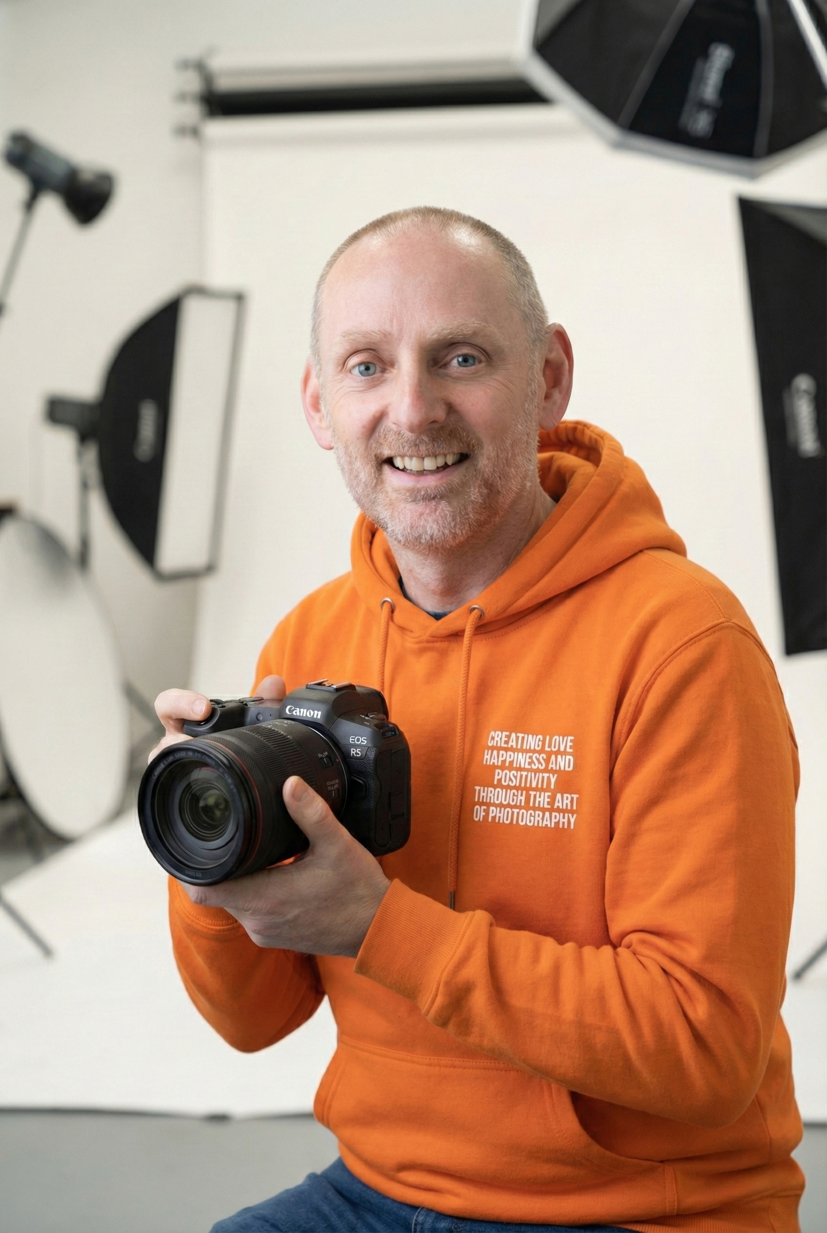 A medium shot of Andrew smiling, sitting in a bright photography studio. He is holding a Canon R5 camera in his hands. He is wearing a vibrant orange hoodie with text printed across the front that reads, 'creating love happiness and positivity through the art of photography.' Professional studio lighting equipment and a white backdrop are visible in the blurred background.