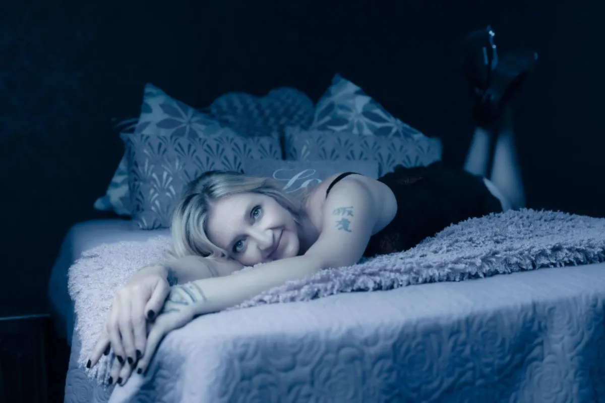 Risbeys Photography: A smiling blonde woman with arm tattoos lying on a bed in our Darlington studio, showcasing the confidence and self-love found through a professional boudoir session in County Durham.