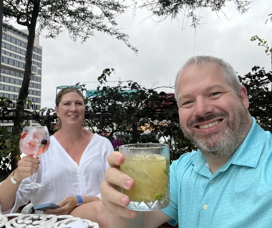 A man and a woman sit at a table holding up cocktails as if toasting the viewer on a rooftop in San Jose, Costa Rica
