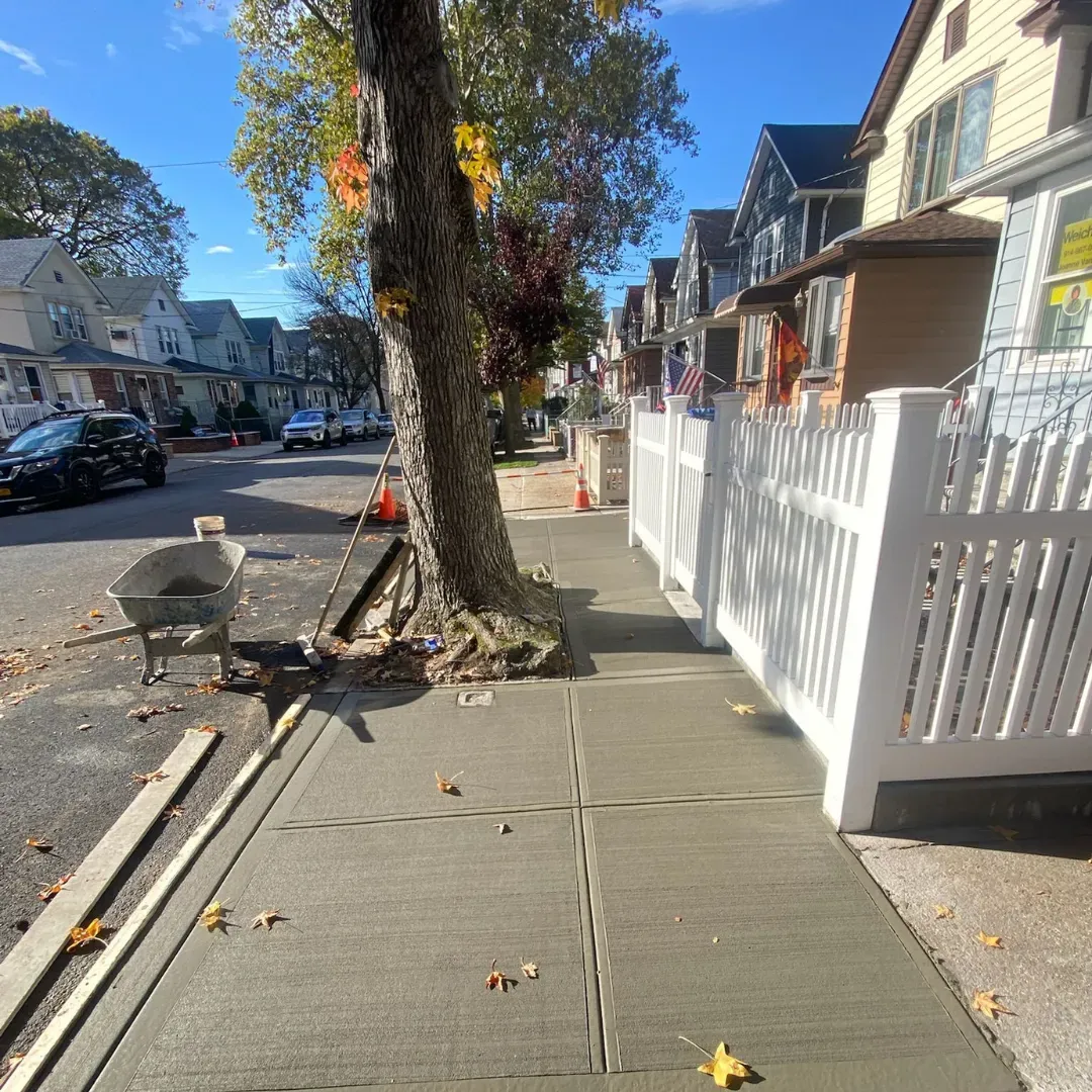 Reconstructed concrete sidewalk in a residential neighborhood of Queens, New York, next to a tree and a white fence.