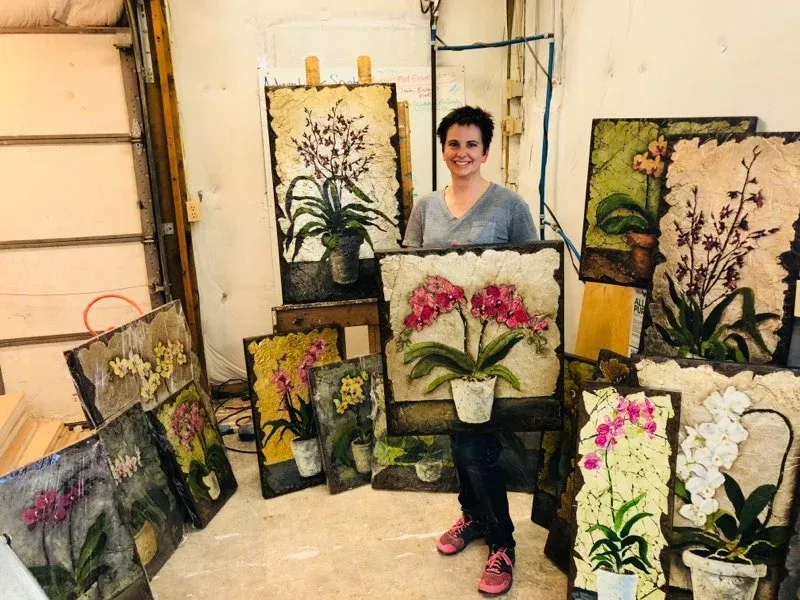 A vibrant, close-up photograph of a blooming orchid with delicate pink and white petals, set against a softly blurred background of an art studio. The image captures the intricate details of the flower and hints of paintbrushes nearby, evoking creativity and serenity.