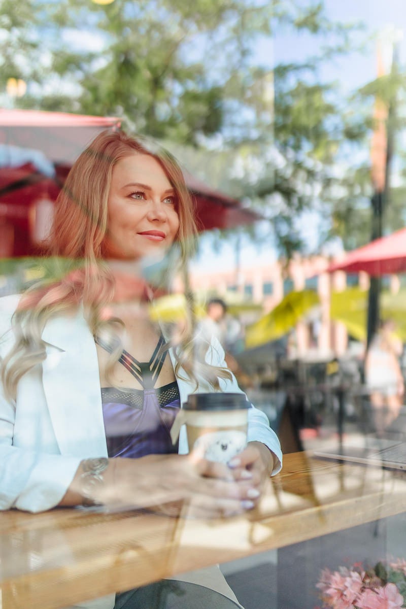 Eva Alexandre, business architecture strategist for established consultants, sitting in a café in Kelowna behind a window with reflections, looking to the side, with a laptop and coffee on the table.