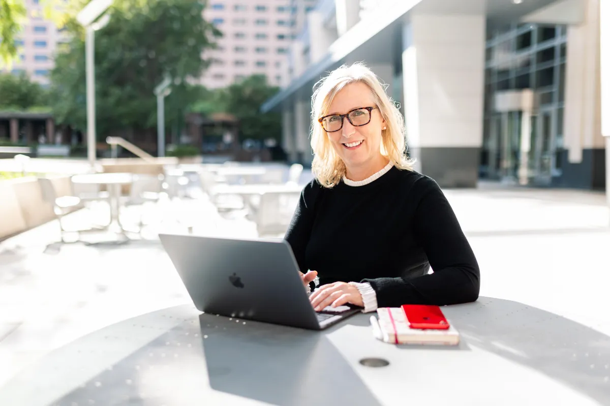 A confident woman in her mid-40s with short hair and a warm, inviting smile sits in a plush armchair. Behind her, a curated bookshelf and soft lighting create a premium, welcoming atmosphere, reflecting wisdom and approachability.
