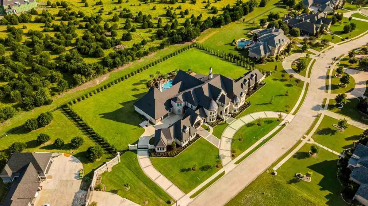 Aerial view of a sprawling mansion surrounded by manicured lawns, circular driveways, and neighboring homes in a suburban setting.