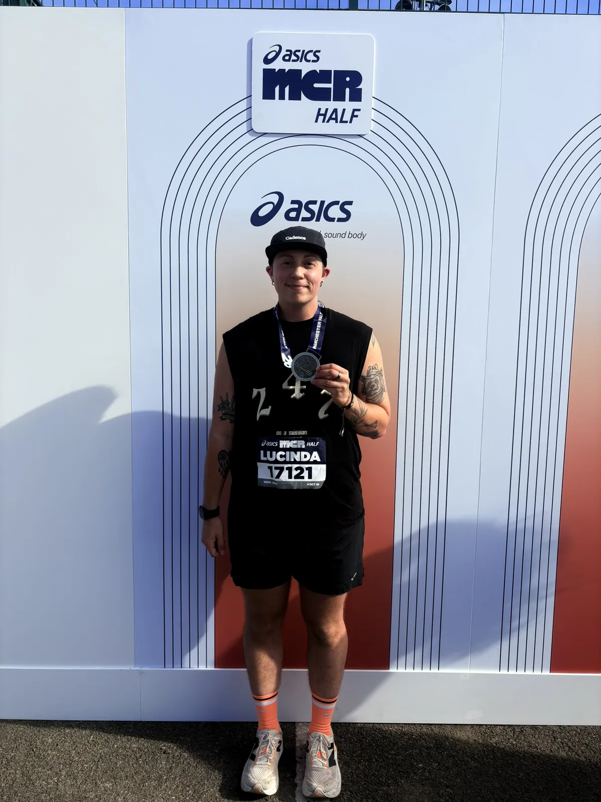 Lucinda Livingstone crossing the finish line of Manchester half marathon, wearing a medal, standing under the Asics arch
