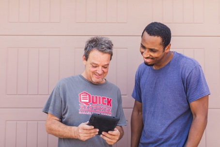 Two men are looking at a tablet and smiling in front of a door.