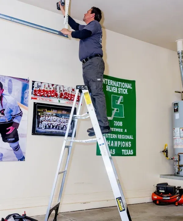 Technician on a ladder installing a garage door opener system inside a residential garage.