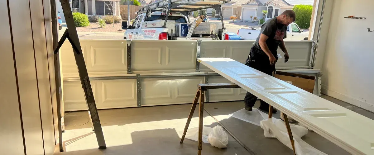 A man in a garage installing a white roll-up door, with tools and a ladder nearby.