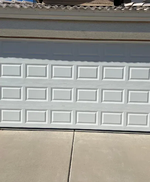 A close-up, straight-on view of a textured white garage door with rectangular panels and light gray concrete driveway.