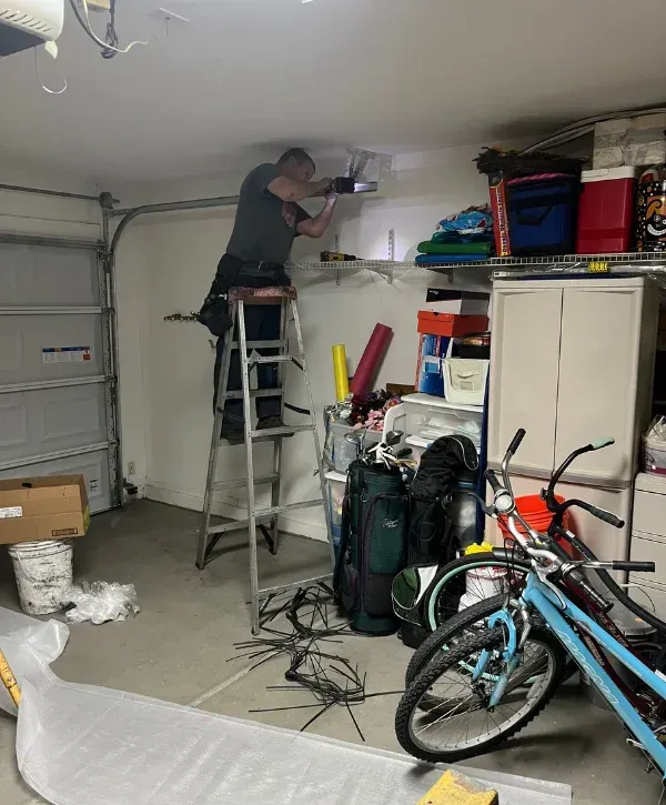 A man on a ladder installs a metal bracket onto a garage wall above shelves, with tools and bicycles nearby.