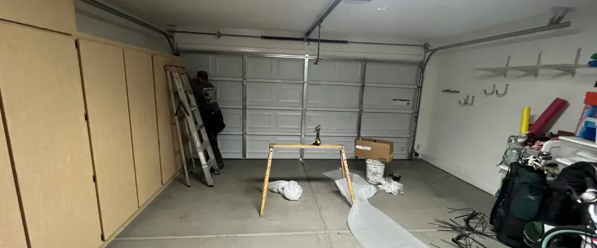 A man on a ladder working on a garage door installation in a messy garage with cabinets, tools, and golf clubs.